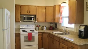 Kitchen with wooden cabinets and granite countertops. White refrigerator, white electric range, stainless steel microwave, and stainless steel sink. Cabinets are wooden with natural finish and silver handles. Countertops are granite with speckled pattern. Red dish towel hanging on oven handle and black coffee maker on counter. Window has red and white checkered curtain. Walls painted light beige color