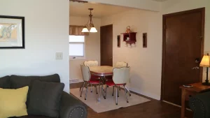 Thank you for sharing the image. Here’s the alt text for the image: “Living room with gray sofa and yellow throw pillow. Wooden side table with lamp next to sofa. Dining area in background with round table and four white chairs around it. Red shelf on wall above table. Walls are white with painting hanging on wall. Floor is wooden with beige rug under dining table. Door on right side of image