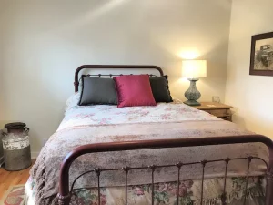 Bedroom with queen size bed with red metal frame. Bed made with white and pink floral comforter and gray and pink throw pillows. Nightstand made of wood with blue lamp on it. Picture hanging on wall above nightstand. Room has hardwood floors and white walls. Metal milk jug on floor next to bed.